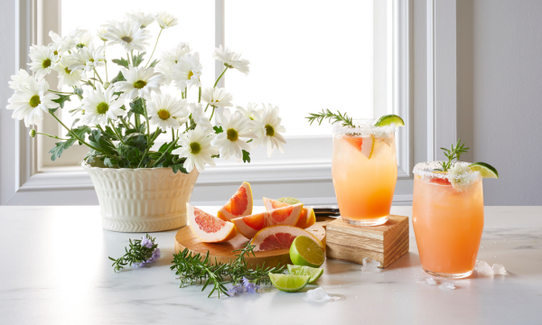 Mocktail Paloma drinks served in glasses and topped with rosemary, grapefruit and lime for garnish. White flowers, and lime wedges, grapefruit, and rosemary ingredients placed next to drinks.
