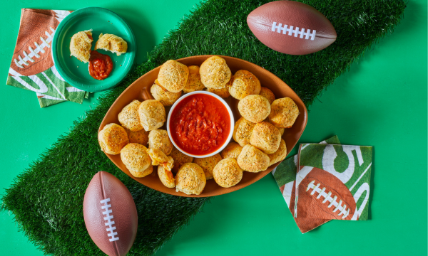 Football shaped platter with pizza rolls and marinara sauce for dipping. Surrounded by footballs, green plates and football napkins.