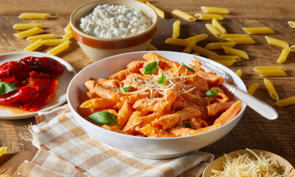 White bowl with penne rigate, topped with shredded parmesan cheese and basil. Surrounded by a bowl of cottage cheese, shredded parmesan cheese, and pasta sauce.