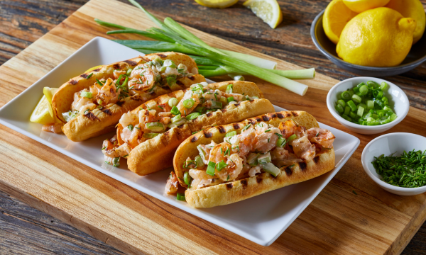 Brioche hot dog buns served with shrimp and topped with green onions and chives. Green onions and lemons on the side, finely chopped ingredients served on white small plates.