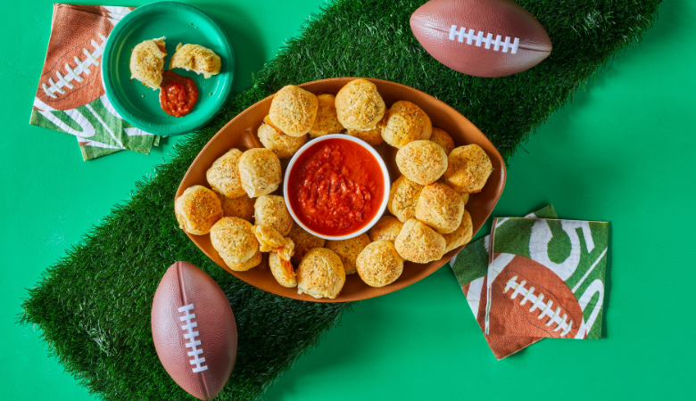 Football shaped platter with pizza rolls and marinara sauce for dipping. Surrounded by footballs, green plates and football napkins.