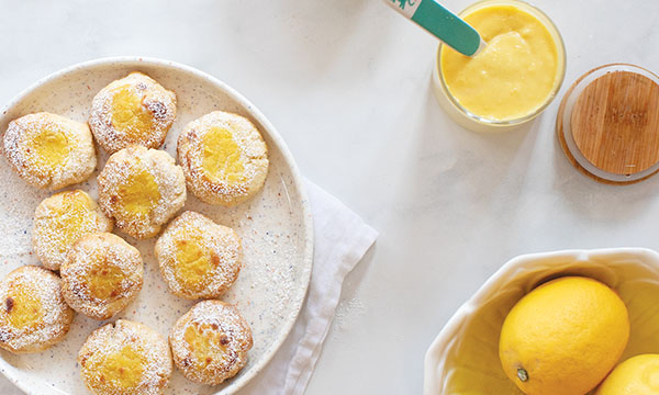 Plate of lemon cookies dusted with powdered sugar, a bowl of fresh lemons, and a jar of lemon curd on a white surface.