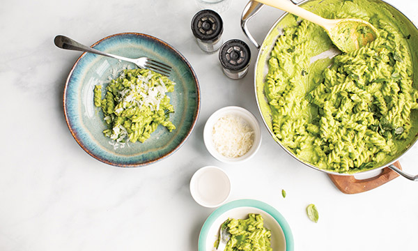 Aerial view of green pasta in a pot, with served portions on plates, topped with cheese, alongside salt and pepper shakers.
