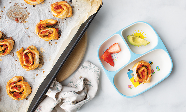 Baked pizza pinwheels on a parchment-lined tray beside a children's divided plate with a pizza roll, sliced strawberries and apple wedges on a light kitchen counter