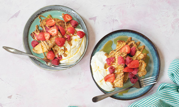 Two blue plates of French toast topped with strawberries and raspberries, served with yoghurt and a drizzle of maple syrup on a light pink surface.