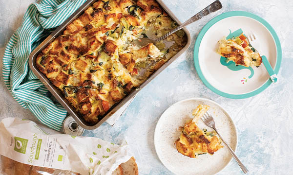 Leek and tomato bread pudding baked in a roasting tin, with portions served on plates beside a loaf of bread and a striped tea towel.