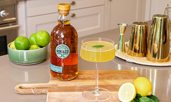 A garnished Sidecar cocktail sits beside a bottle of Roe & Co. whiskey on a wooden board. Nearby are limes, a lemon, mint leaves, and gold cocktail shakers.