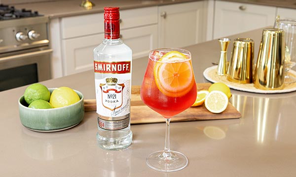 A bottle of vodka beside a vibrant red-orange cocktail garnished with lemon slices. Nearby, a bowl of citrus fruits and gold cocktail shakers create a fresh, elegant vibe.