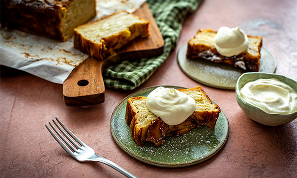Slices of invisible apple cake topped with cream on green ceramic plates, with a loaf on a wooden board in the background.