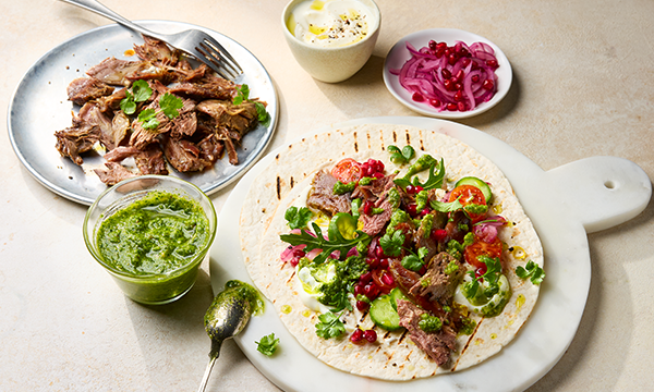 A vibrant wrap topped with sliced lamb leftover lamb from Easter, fresh greens, tomatoes, pomegranate seeds, and salsa verde sauce on a white board. Nearby, a plate with lamb and salsa verde sauce..