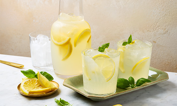 Chilled lemon basil punch drink with lemon slices and basil garnishes in glasses on a tray, accompanied by a pitcher and a plate of lemons on a marble surface.