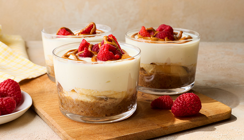 Three glass cups of cheesecake breakfast pots sit on a wooden board, topped with fresh raspberries and caramel drizzle. A small plate with raspberries is nearby.