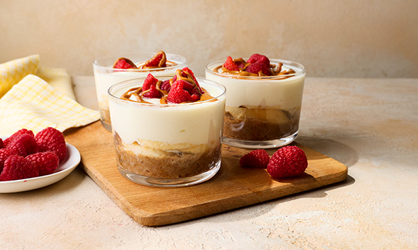 Three glass cups of cheesecake breakfast pots sit on a wooden board, topped with fresh raspberries and caramel drizzle. A small plate with raspberries is nearby.
