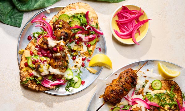 Grilled chicken shawarma on flatbread with sliced cucumber, pickled onions, and pomegranate seeds. A lemon wedge and extra onions are beside them.