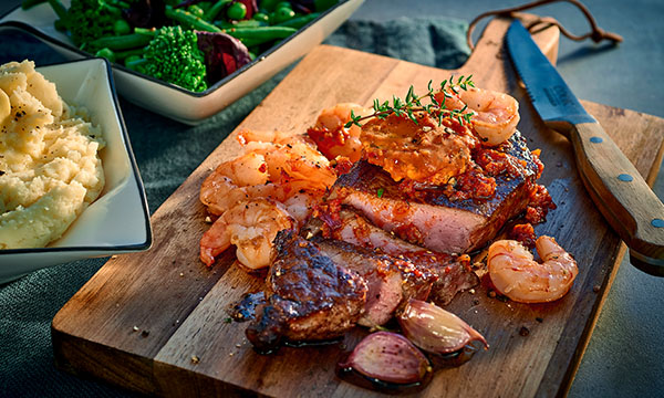 Grilled steak topped with seasoned shrimp, roasted garlic, and herbs, served on a wooden board with sides of mashed potatoes and steamed vegetables.