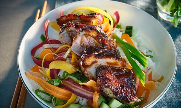 A bowl of rice topped with glazed grilled duck and colourful fresh vegetables, including bell peppers, carrots, and cucumbers.