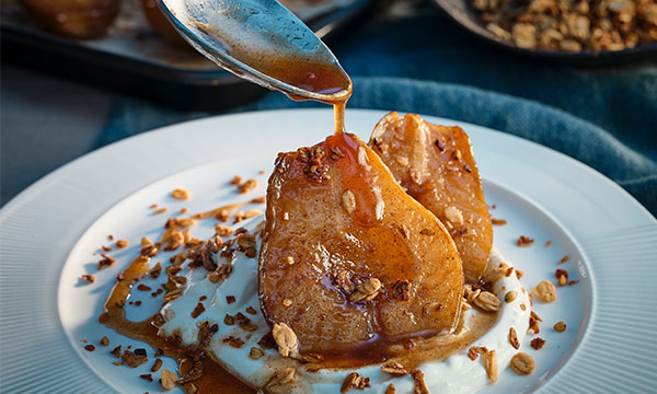 Caramel-drizzled poached pears on creamy yogurt, topped with crunchy granola, served on a white plate with a blue background.