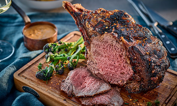 Juicy, medium-rare prime rib roast on a wooden board with grilled broccolini and a side of creamy sauce.