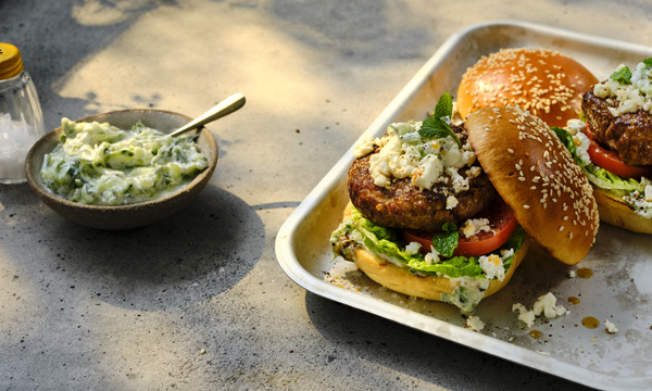 Two gourmet burgers with lettuce, tomato, crumbled feta cheese, and creamy Tzatziki on sesame brioche buns