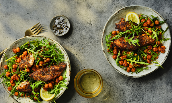 Two plates of seasoned grilled chicken served over green leaves, roasted chickpeas, and lemon wedges