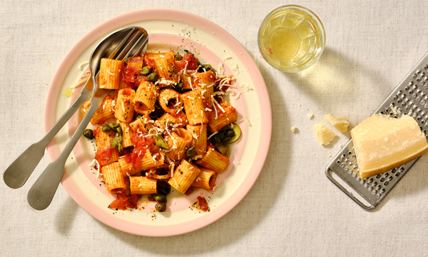 a bowl of penne pasta in a tomato based sauce, capers, blacks olives and anchovies