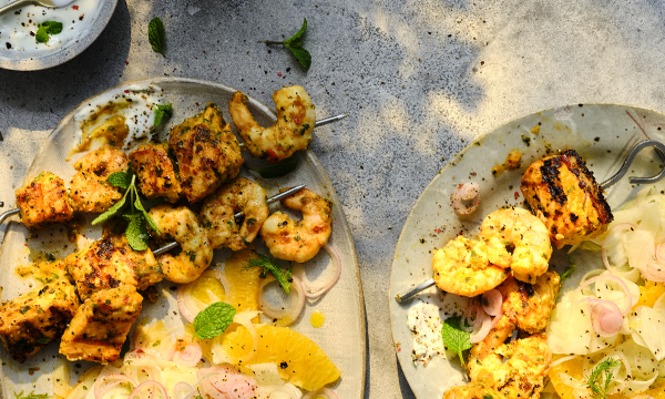Grilled skewers of prawns and salmon served on plates with slices of orange, shallots, herbs, and a lime yogurt