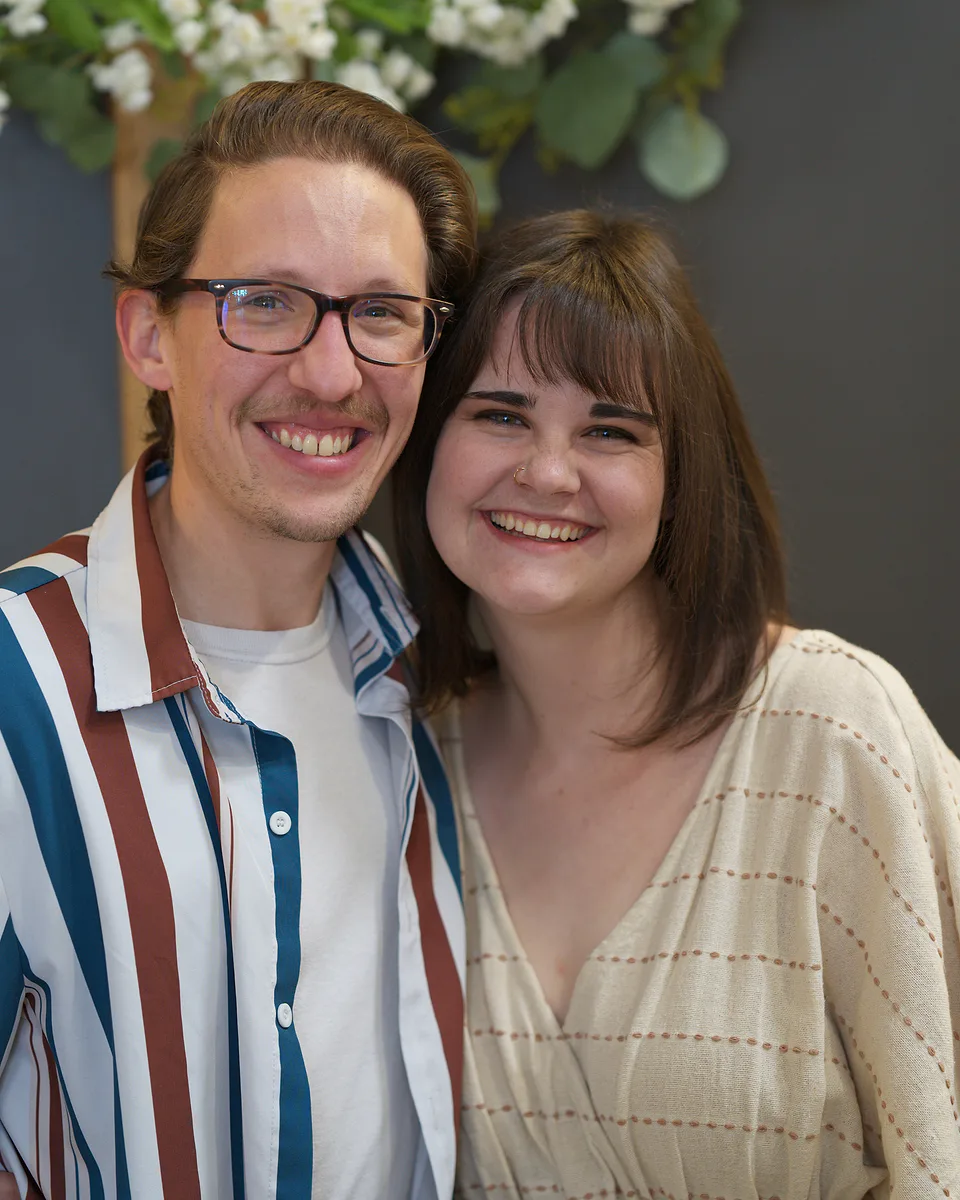 A couple smiling cheek to cheek against a grey wall, he in a striped shirt and glasses, she in a cream knit top, white flowers above them