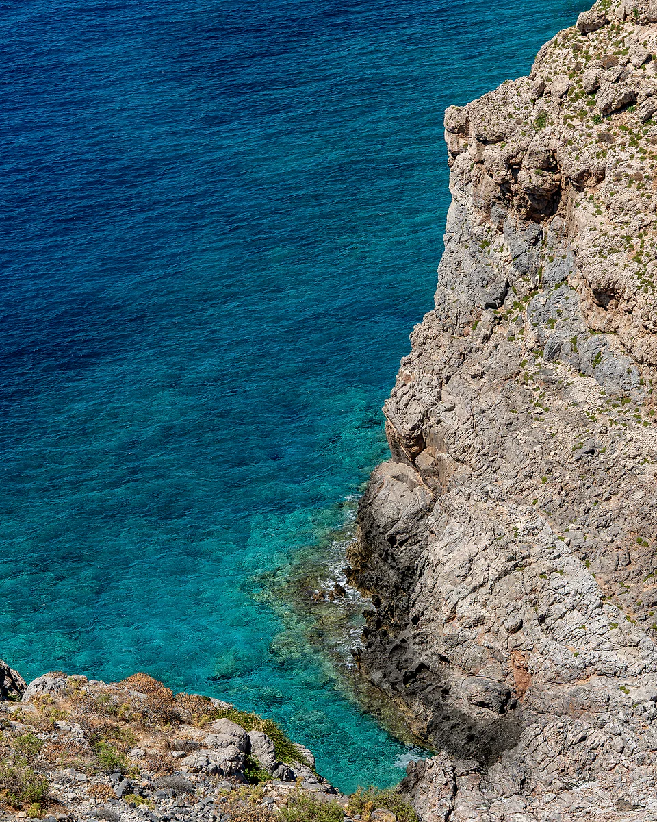 Rocky cliff seen from above, dropping into turquoise water that deepens to dark blue