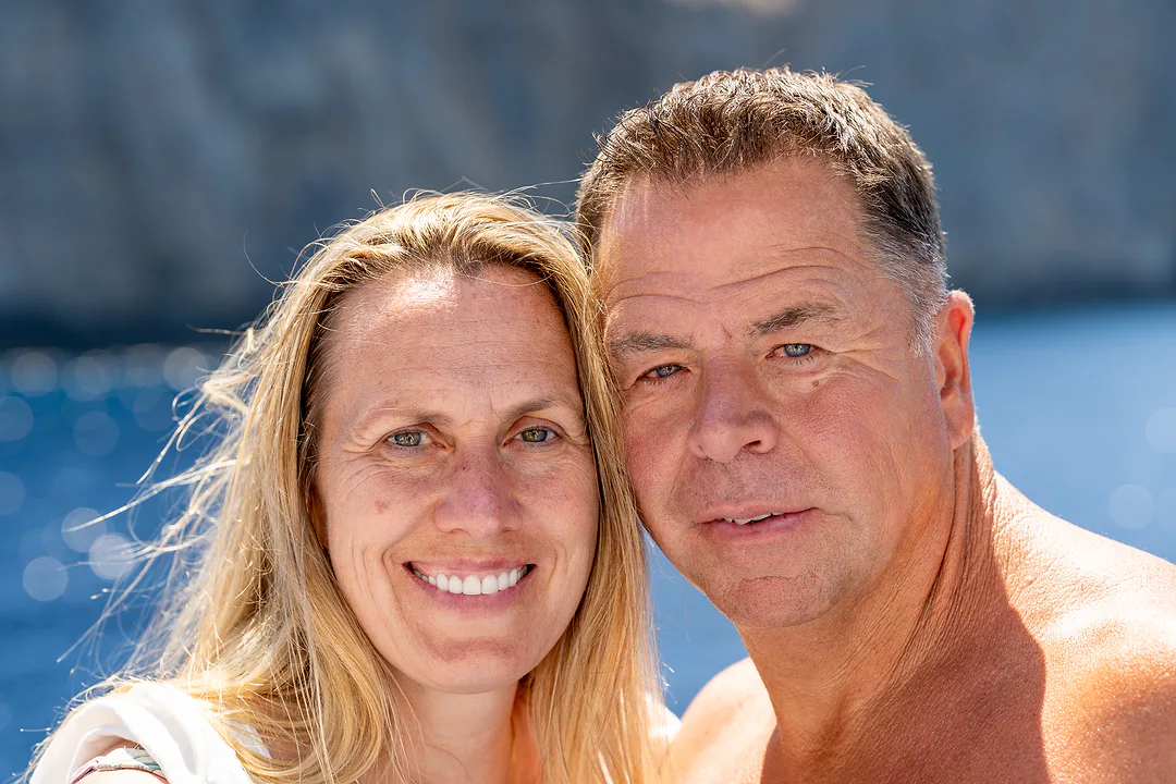 A couple cheek to cheek and smiling in bright sun, sunlit water sparkling behind them