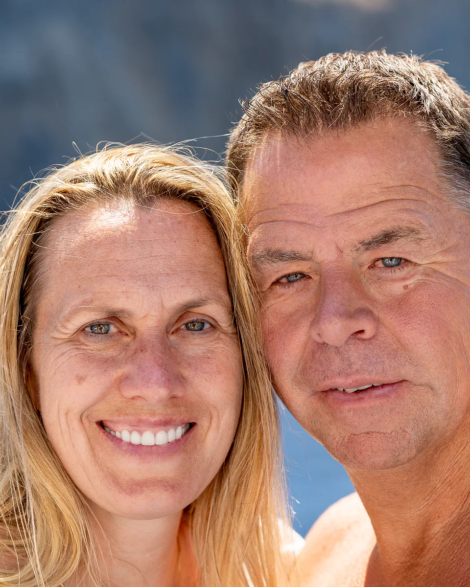 A couple cheek to cheek and smiling in bright sun, sunlit water sparkling behind them