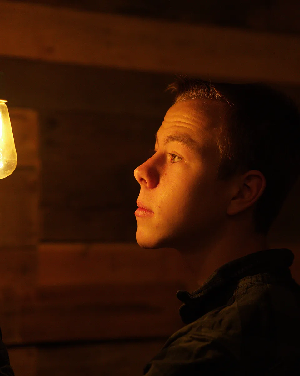 Young man in profile lit only by a hanging Edison bulb, one hand raised toward it against dark barn wood