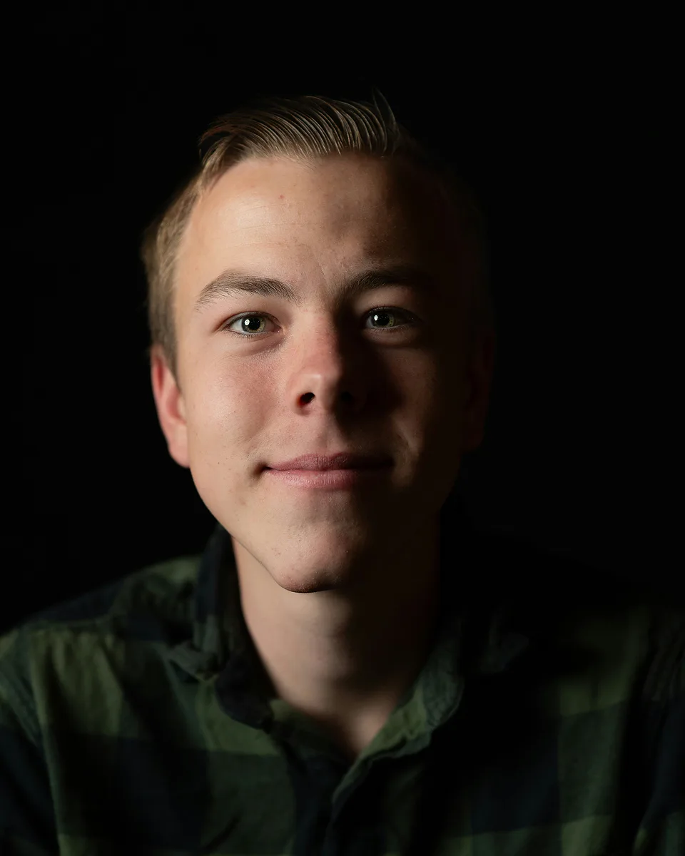 Low-key studio portrait of a young man in a dark green plaid shirt, one side of his face lit against a black background