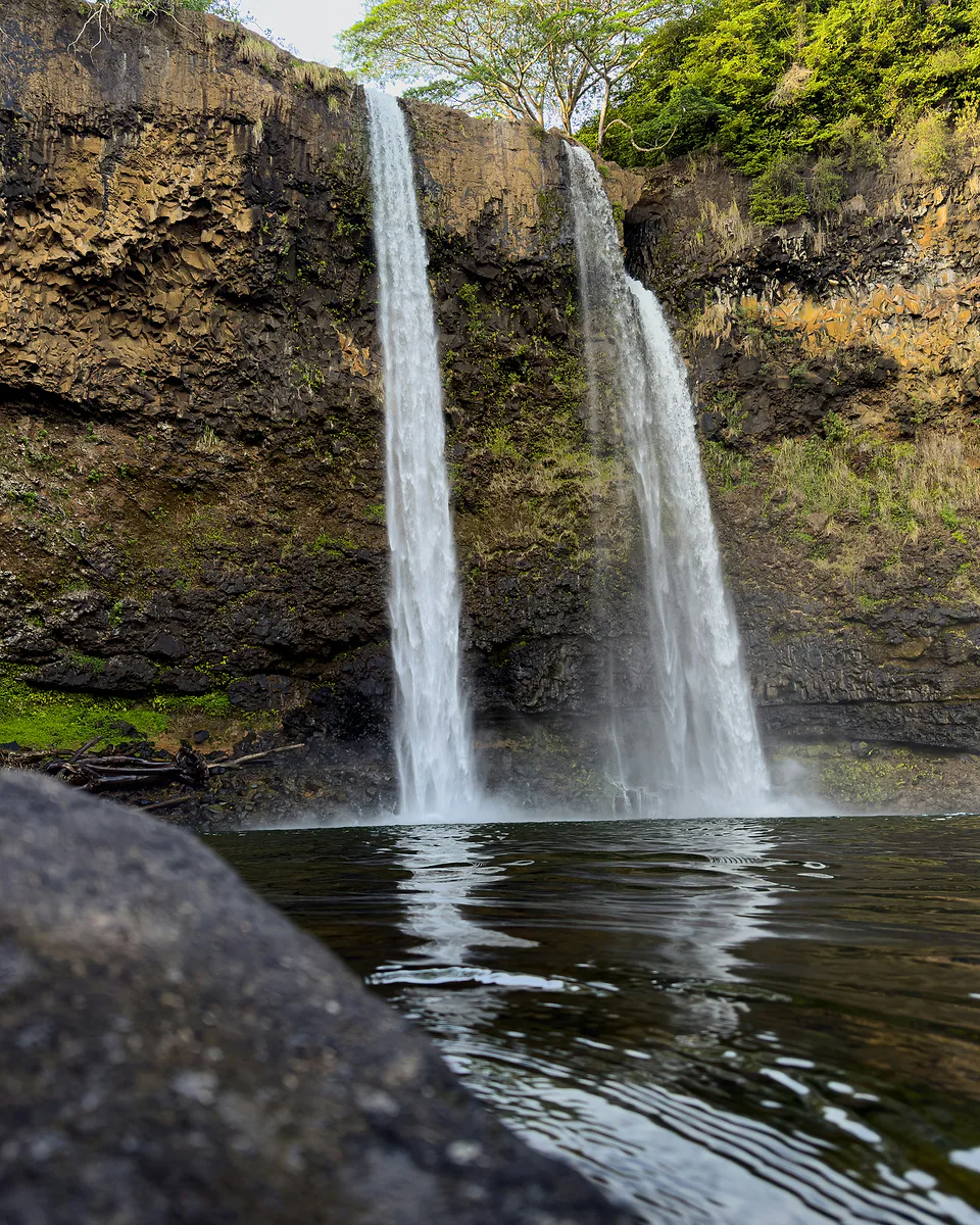 Twin waterfalls dropping side by side over a mossy cliff into a calm dark pool, seen from a rock at the water's edge