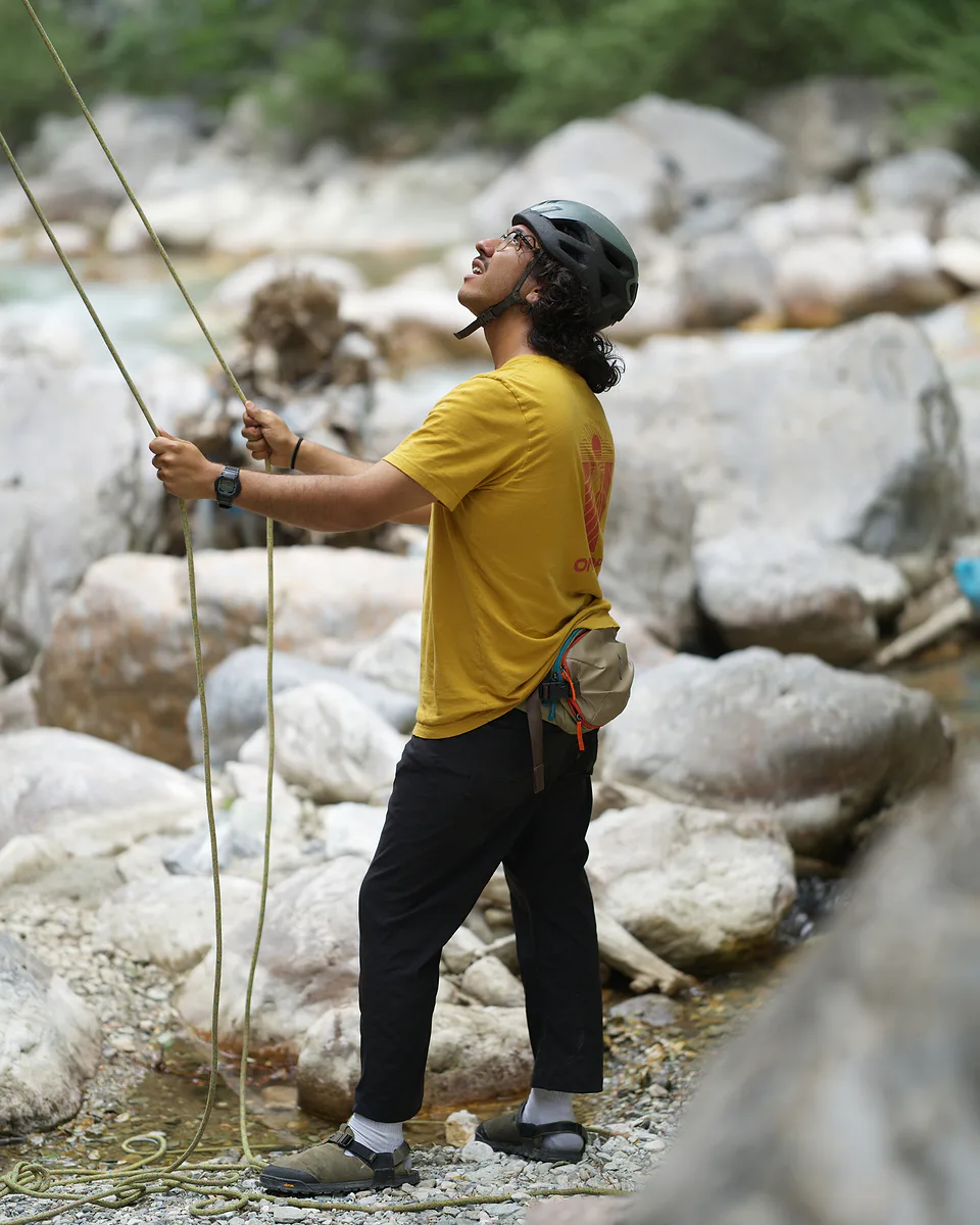 Man in a yellow shirt and climbing helmet feeding rope through his hands and looking up, standing on pale boulders beside a canyon river