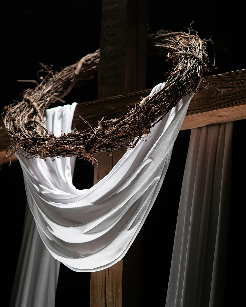 A wooden cross draped with white cloth and a crown of thorns, lit against a black background