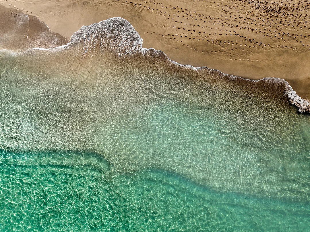 Aerial view of clear turquoise shallows meeting a tan beach, a line of white foam and footprints across the wet sand