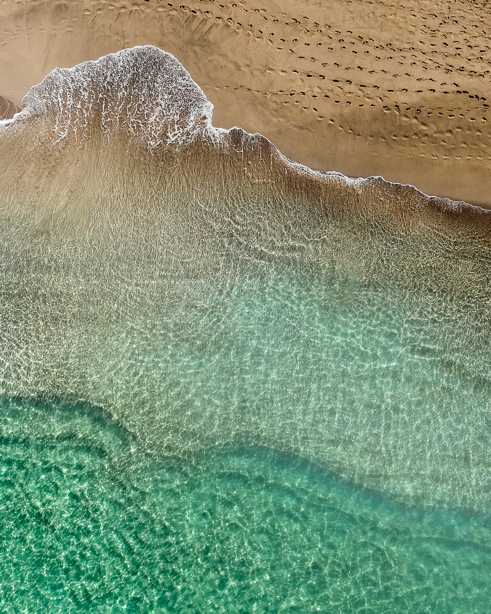 Aerial view of clear turquoise shallows meeting a tan beach, a line of white foam and footprints across the wet sand