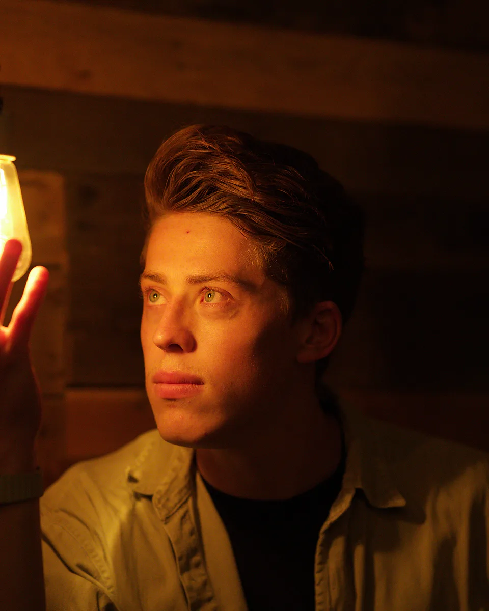 Young man in a dim wood-panelled room holding his hand beside a hanging Edison bulb, its warm light glowing through his fingers