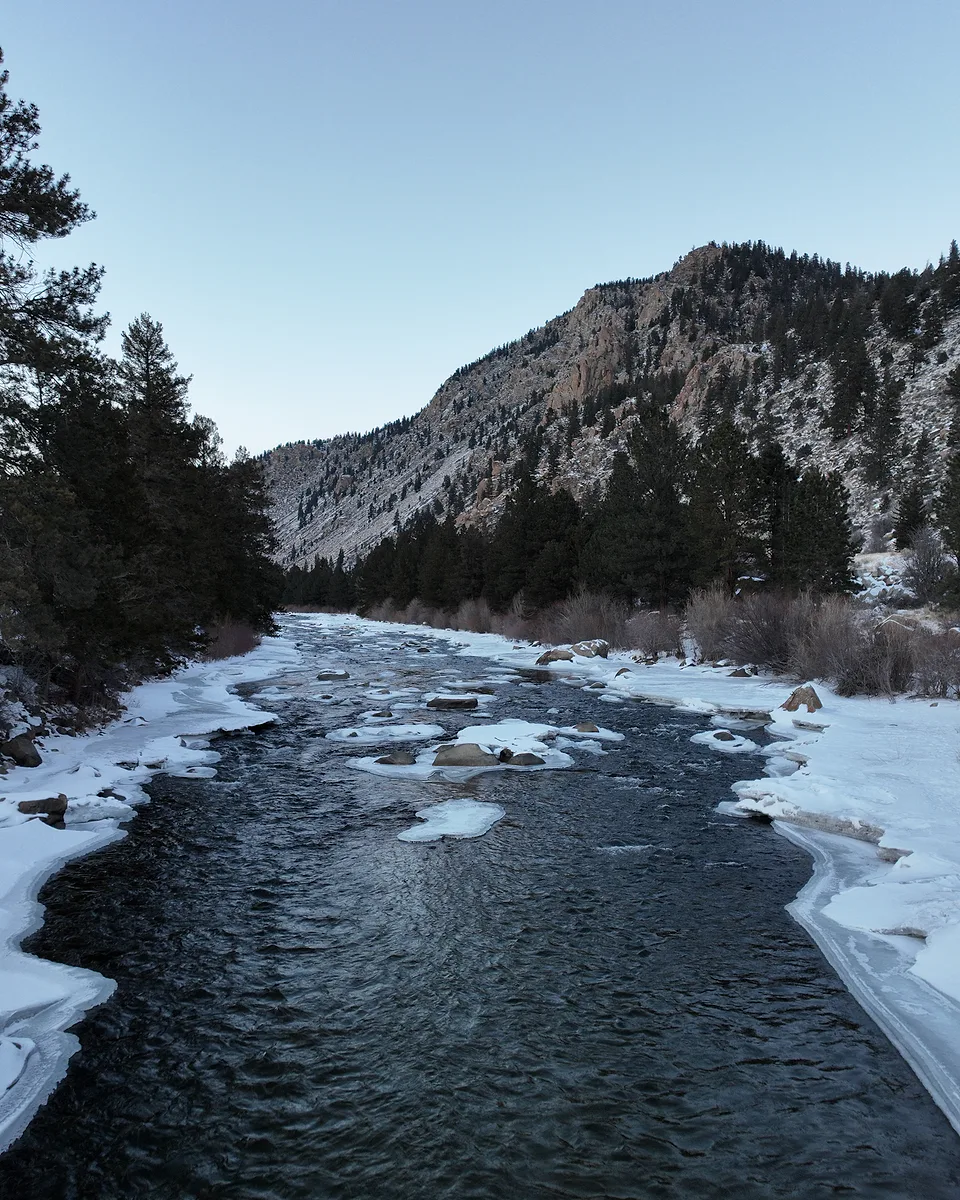 A dark river running between snow-covered banks in a pine valley, a rocky ridge catching the last of the light