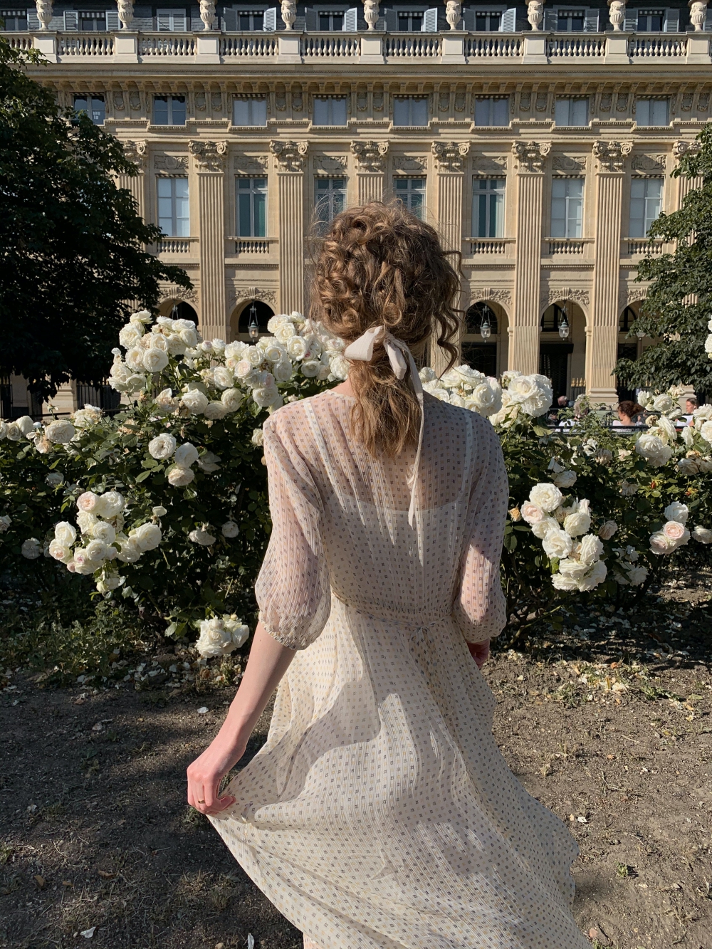 A girl in a white dress walking towards a rose bush in front of a white building