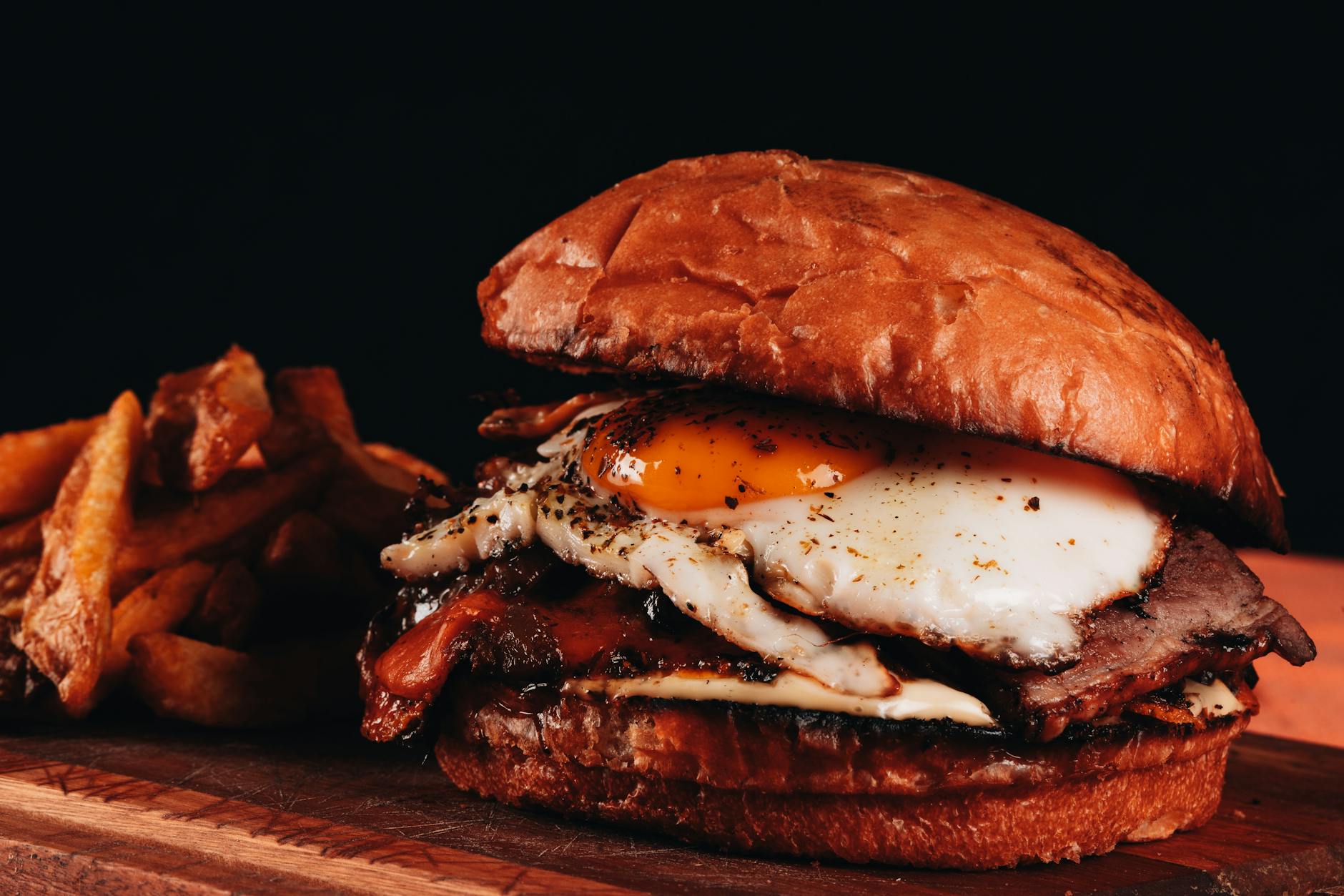 Close-up of a gourmet burger with fried egg, bacon, and fries on a wooden board.