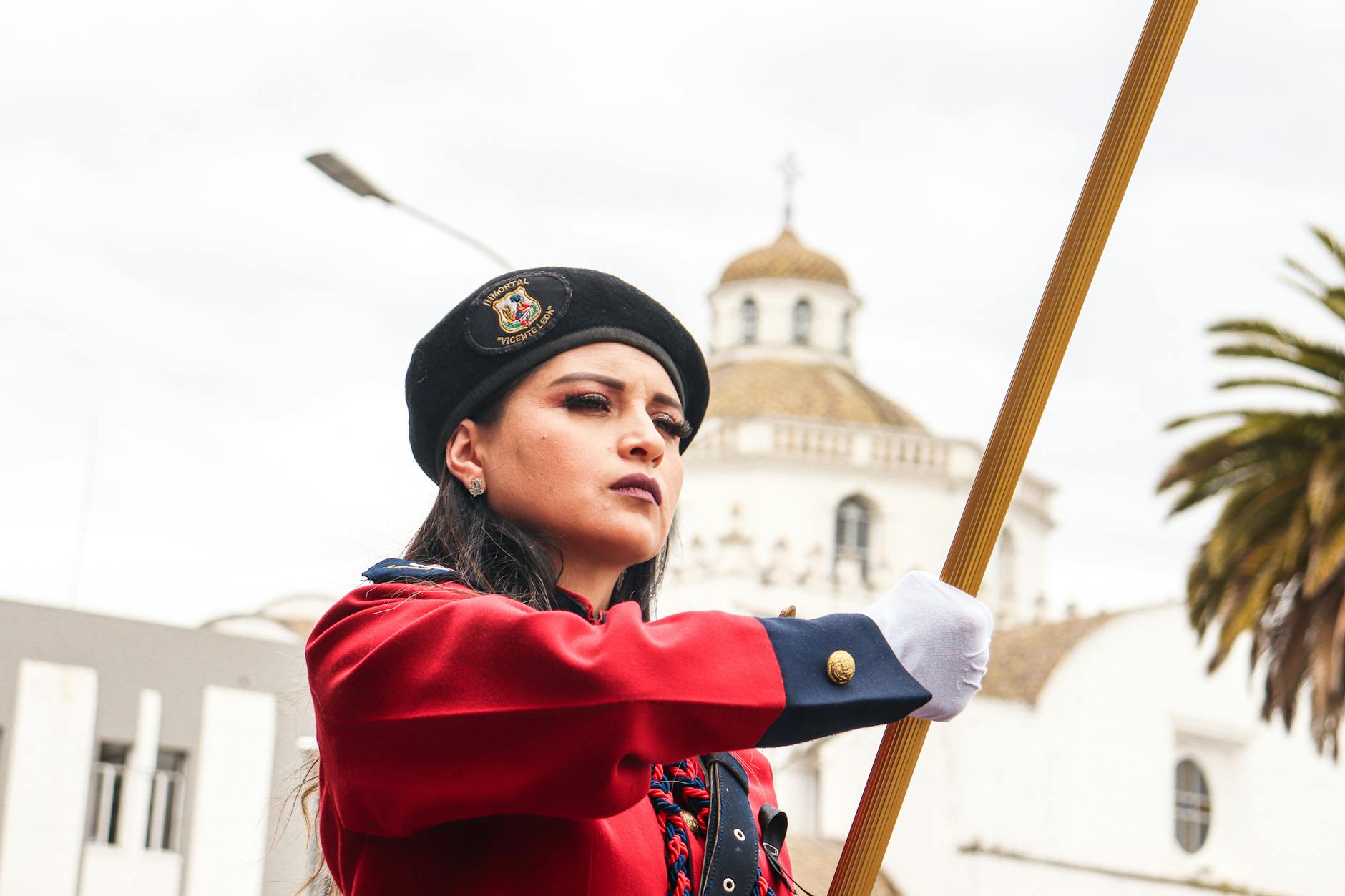 Young woman in military-style uniform holding a flag during a parade in Ecuador, with a historic building in the background.