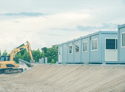 Graue Containeranlage mit einem gelben Raupenbagger links, im Hintergrund Bäume & Sträucher