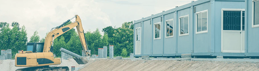 Graue Containeranlage mit einem gelben Raupenbagger links, im Hintergrund Bäume & Sträucher