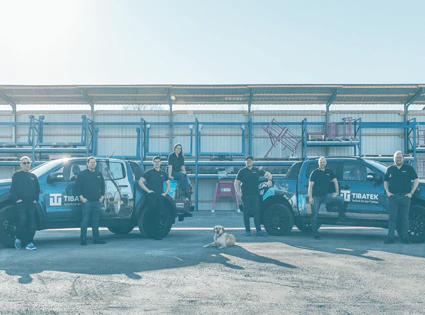 Tibatek Teamfoto mit sieben Personen vor zwei gebrandeten Pickups und einem Hund