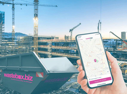 Hand hält Smartphone mit wastebox.biz App vor einer großen Baustelle im Hintergrund