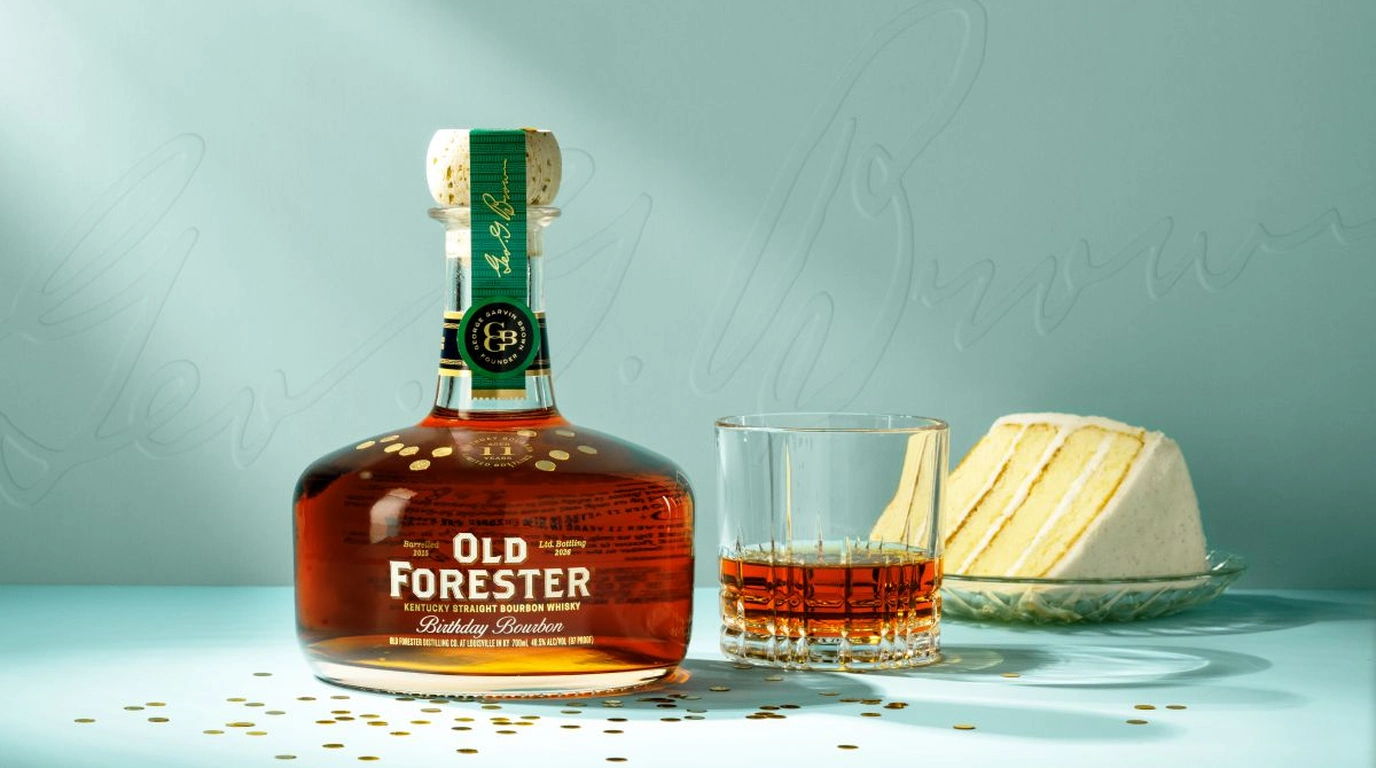 Old Forester 2026 Birthday Bourbon bottle with its green George Garvin Brown neck ribbon beside a rocks glass pour and a slice of white layer cake on a pale blue backdrop