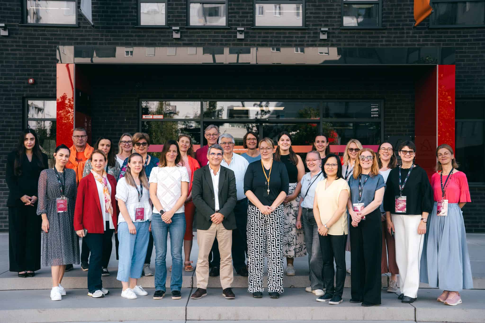 A diverse group of 24 people stand in front of a modern building with a black facade and red accents, smiling warmly, conveying a sense of camaraderie.