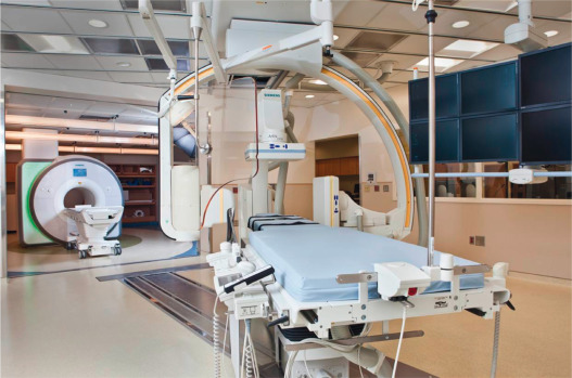 FIG. 47.1, National Heart Lung and Blood Institute/Children's National Medical Center combined x-ray and interventional cardiovascular magnetic resonance (iCMR) suite, Washington, DC. The iCMR suite combines a biplane x-ray system (foreground) and a 1.5 T CMR scanner (background). The x-ray table docks with the CMR table to enable the patient to be easily transferred between the two modalities.