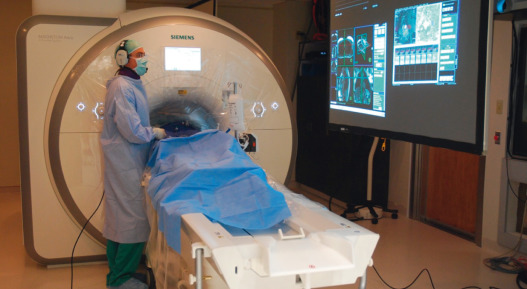 FIG. 47.2, Working inside the interventional cardiovascular magnetic resonance (iCMR) catheterization laboratory. The operator communicates with the patient and with staff inside and outside the iCMR room with sound-suppression microphones and headsets. Real-time CMR, scanner control interface and patient hemodynamics are displayed inside the room using video projectors.
